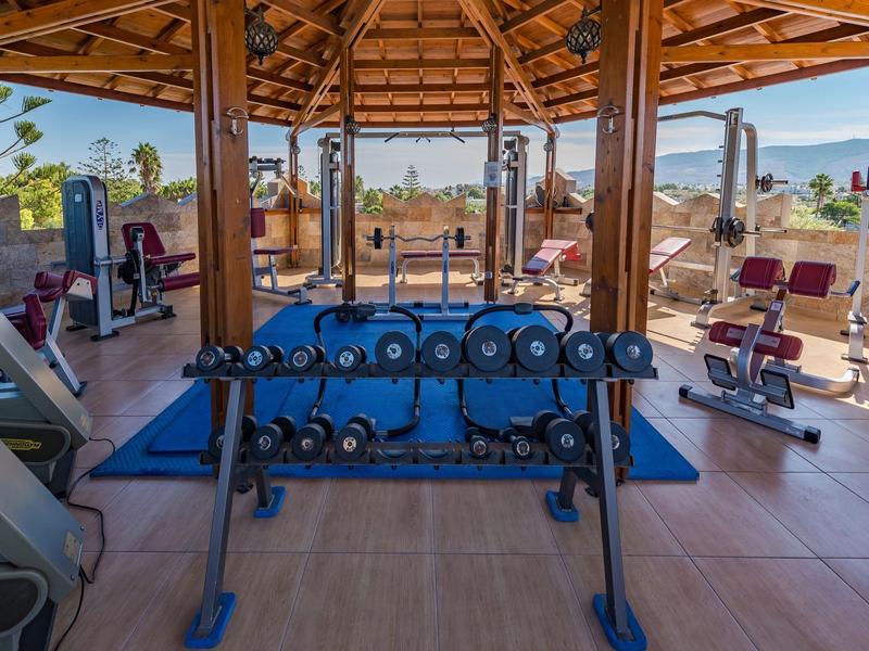 Siłownia na zewnątrz hotelu z ciężarami, maszynami do ćwiczeń i widokiem na ocean pod drewnianym dachem.
