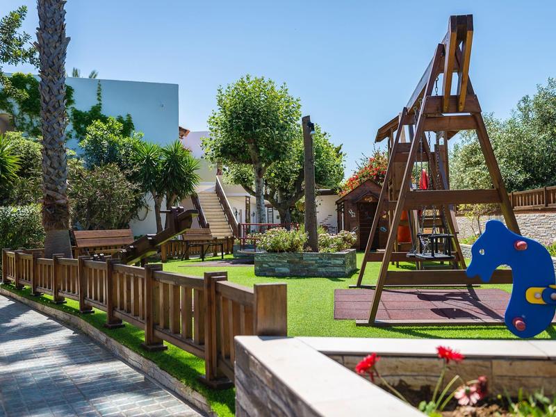 Słoneczny plac zabaw z huśtawkami i miejscami do siedzenia otoczony zielenią i budynkami.