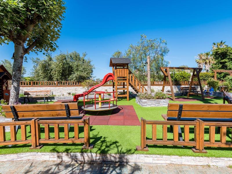 Słoneczny plac zabaw z drewnianymi ławkami, zjeżdżalnią, huśtawkami i zielonym trawnikiem pod jasnym niebem.