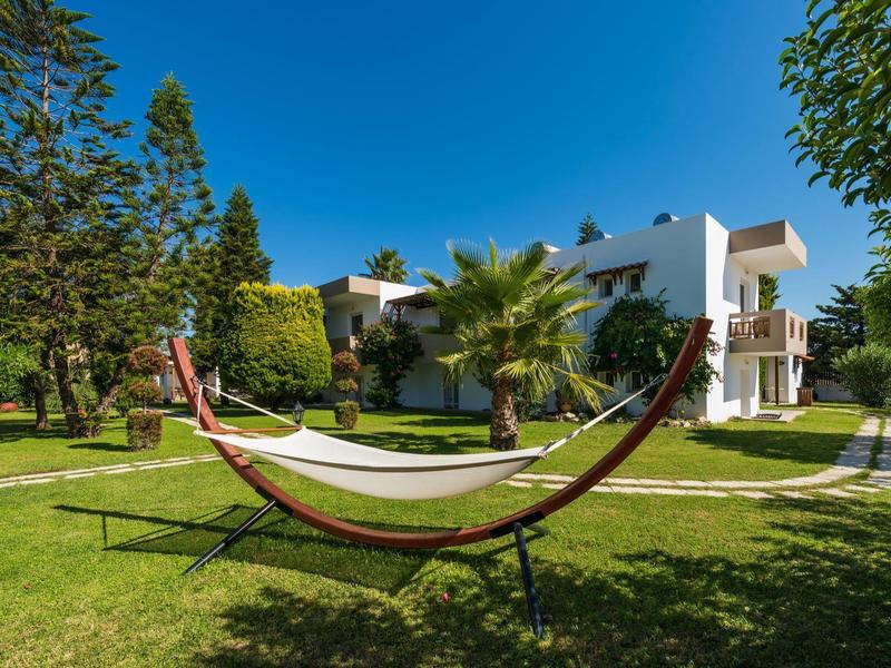 Hamak na stelażu na trawniku z drzewami i białym budynkiem hotelu pod jasnym niebieskim niebem.