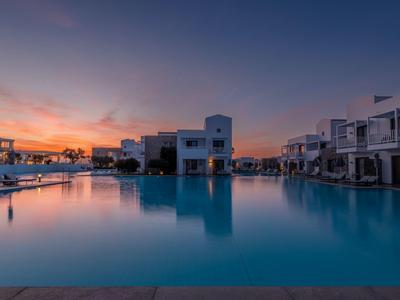 Large hotel pool at sunset with modern white buildings in the background.