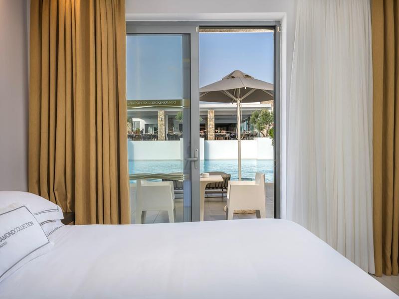 Bedroom with white bed and curtains overlooking a pool and sun umbrella.