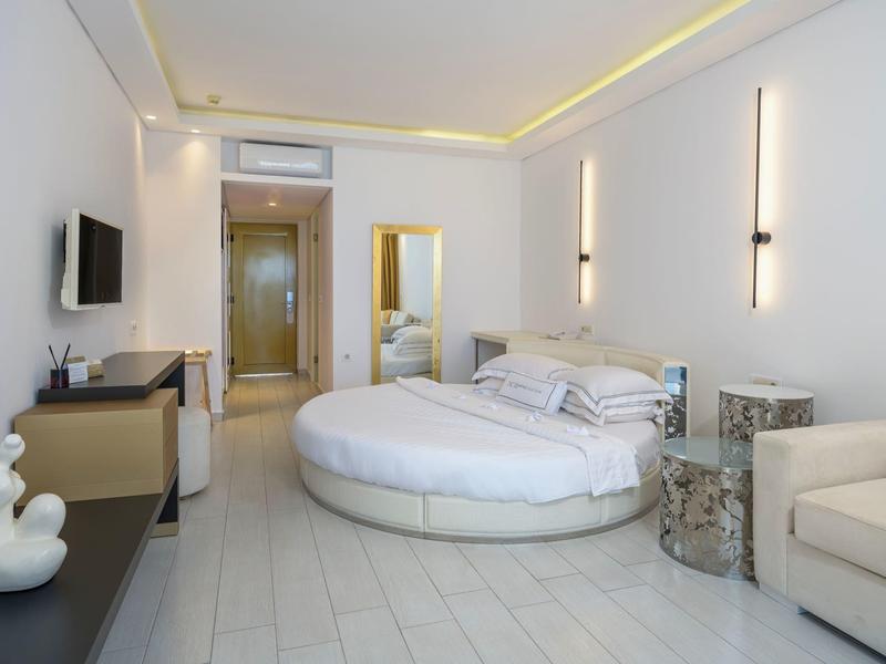 Modern hotel room with round bed, sofa, TV, and minimalist white design.