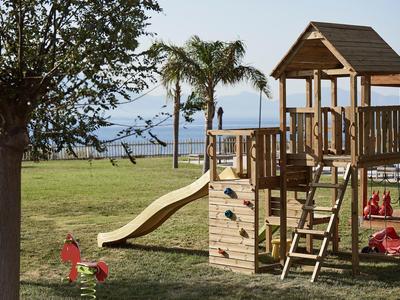 Spielplatz mit Holzklettergerüst, Rutsche und Sandkasten auf einer grünen Wiese am Meer.