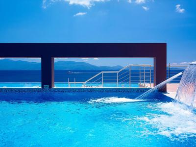 Blauer Pool mit Wasserfall, moderner Rand und Meer mit Bergen im Hintergrund bei klarem Himmel.