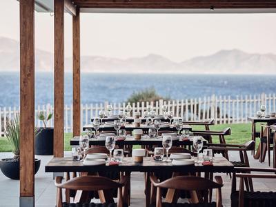 Restaurantterrasse mit Holztischen und Stühlen, Meerblick und Bergen im Hintergrund.