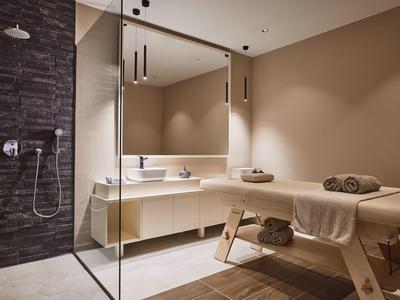 Moderner Spa-Raum mit Massageliege, Handtüchern, großem Spiegel und Duschbereich mit dunklen Fliesen.