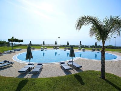 Runder Pool mit Liegen, Sonnenschirmen und Palmen, davor grüner Rasen, im Hintergrund Meer.