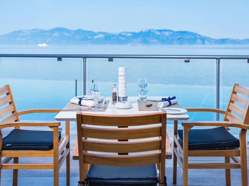 Zona pranzo moderna con mobili in legno su balcone con vista sul mare e montagne.