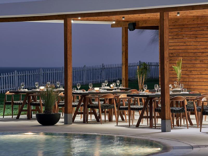 Area ristorante all'aperto con mobili in legno e vista mare al tramonto.