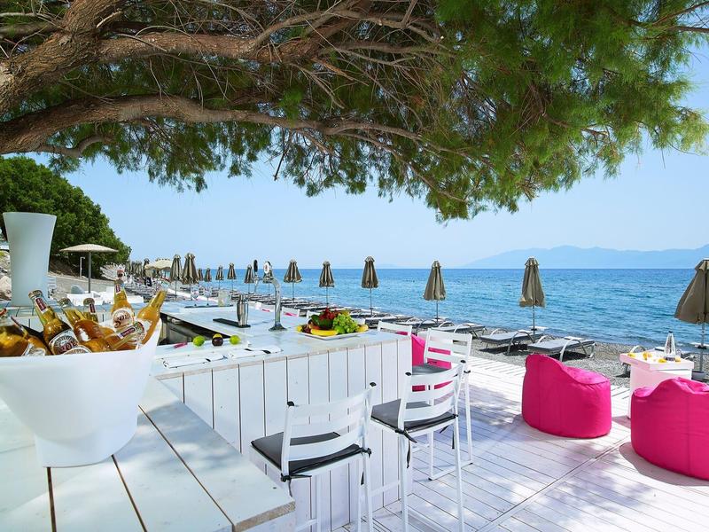 Area spiaggia moderna con mobili bianchi, pouf rosa e vista sul mare sotto un cielo soleggiato.