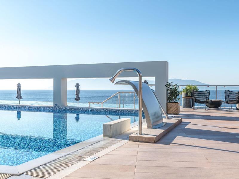 Piscina moderna con scivolo in acciaio inox e area salotto con vista sul mare sotto un cielo limpido.