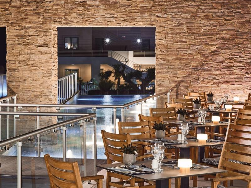 Ristorante all'aperto con tavoli e sedie in legno davanti a muro in pietra, vista sulla piscina illuminata.