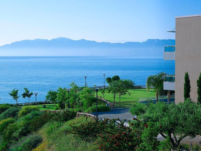 Vista sul mare con scogliere, cespugli verdi e una parte di edificio sotto un cielo limpido.