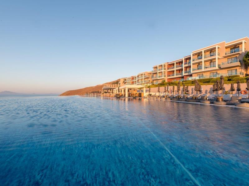 Complesso alberghiero moderno con piscina a sfioro sul mare sotto un cielo limpido al tramonto.