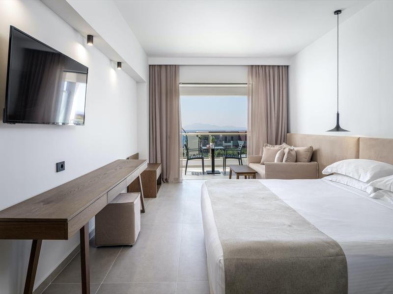 Camera d'hotel luminosa con letto grande, divano, scrivania e balcone con vista mare.