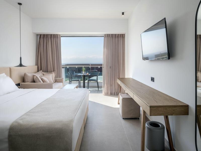 Camera d'hotel moderna con letto matrimoniale, divano, TV a schermo piatto e balcone con vista mare.