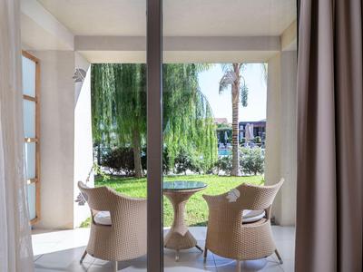 View through glass doors to a balcony with two chairs and a table, beyond a green garden.