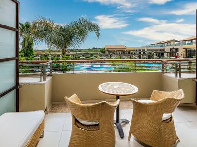 Balcony with table and two chairs overlooking pool and hotel buildings under blue sky.