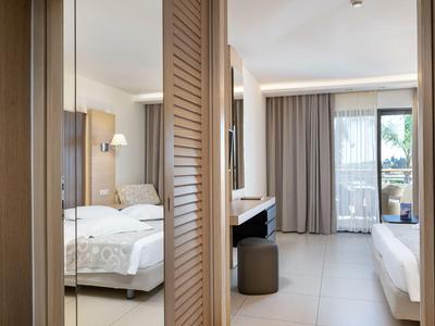 View of a bright hotel room with beds, desk, and balcony with curtains.