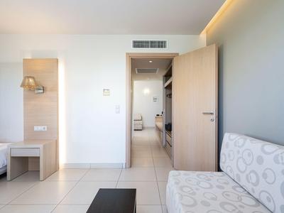 Bright hotel room entrance with open wooden door, sofa, and visible bed.