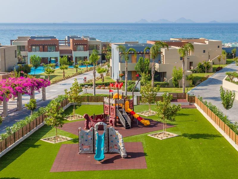 Moderner Wohnkomplex mit Spielplatz, grünen Rasenflächen und Meerblick im Hintergrund.