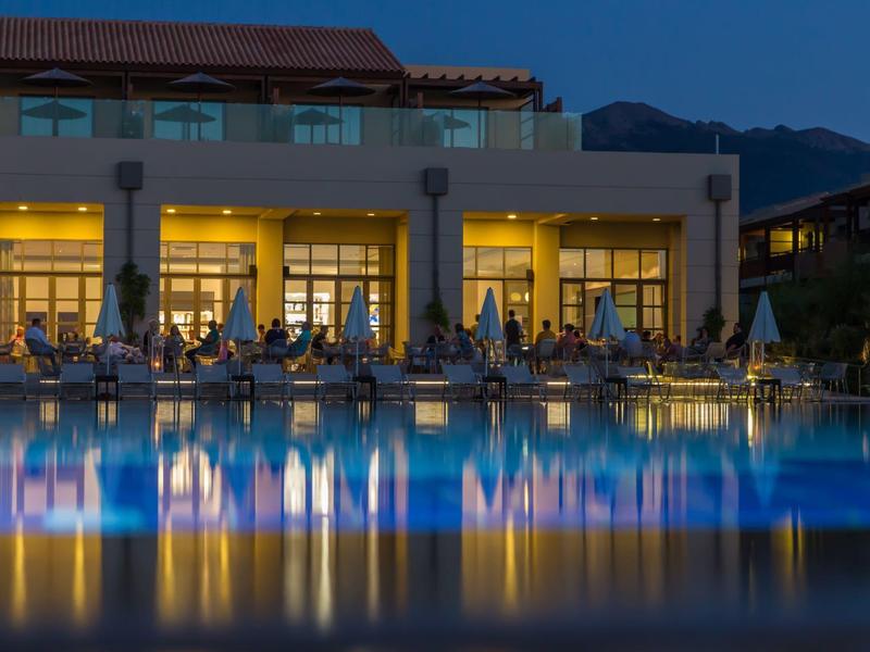 Beleuchteter Pool vor Hotelgebäude in der Abenddämmerung mit Bergkulisse.