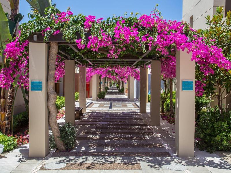 Blumenüberwachsener Pergolaweg mit rosa Blüten in einem Hotelgarten bei klarem Himmel.