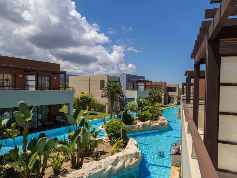 Blick auf luxuriöse Hotelanlage mit Pool, tropischen Pflanzen und modernen Gebäuden unter blauem Himmel.