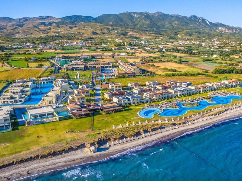 Luftaufnahme eines Strandresorts mit Pools, Gebäuden und Bergen im Hintergrund.