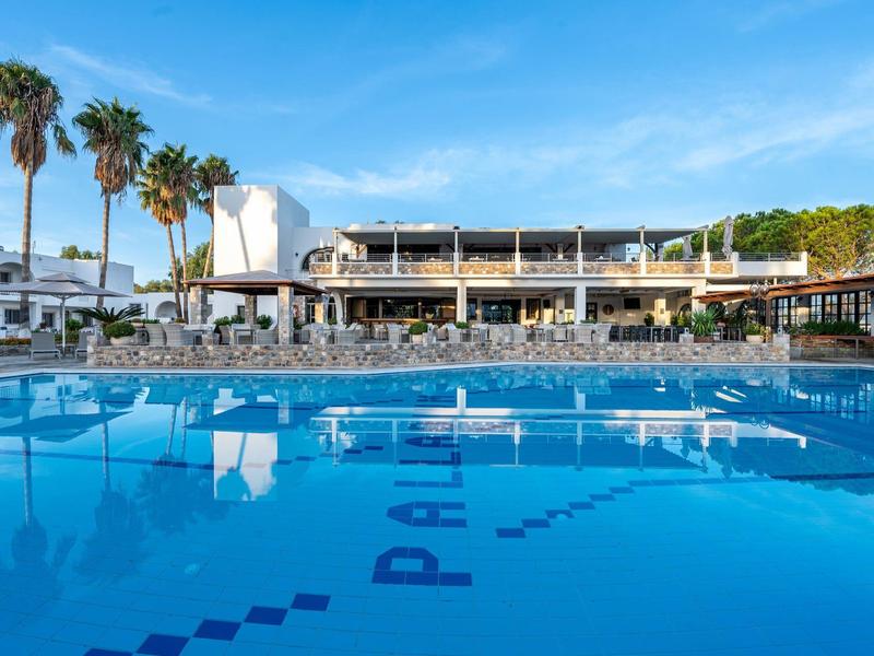 Piscine claire devant un hôtel moderne avec des palmiers et un ciel bleu.