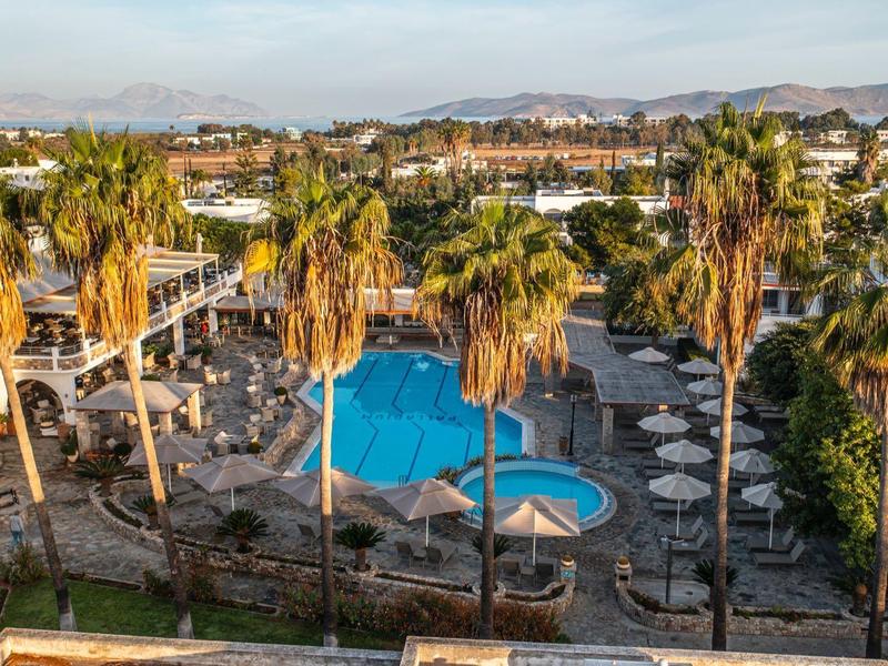 Vue sur une piscine d'hôtel avec palmiers et parasols sous un ciel ensoleillé.