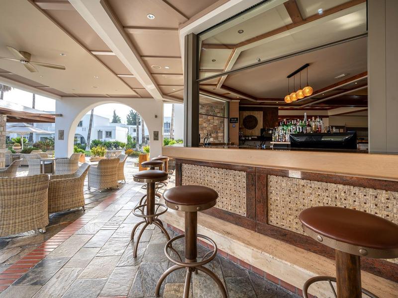 Bar d'hôtel ouverte avec tabourets marron et fauteuils en rotin sur sol carrelé.