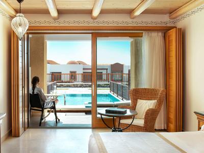 Camera d'hotel con balcone che si affaccia su una grande piscina all'aperto sotto un cielo sereno.