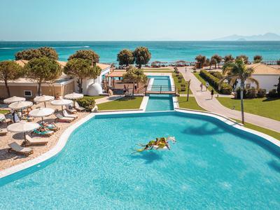Blick auf einen großen Pool mit Liegestühlen und Meer im Hintergrund bei klarem Himmel.