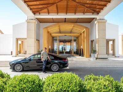 Uomo accanto a un'auto nera davanti all'ingresso di un hotel moderno con tetto in legno e porte di vetro.