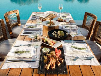 Une table en bois dressée au bord de la piscine avec un plat de fruits de mer et des verres à vin.