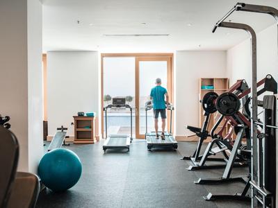 Salle de sport avec équipements d'entraînement et une personne sur un tapis roulant près de grandes fenêtres.