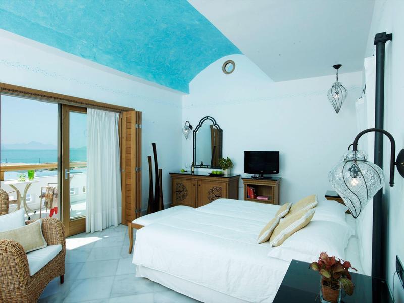 Camera d'hotel luminosa con soffitto turchese inclinato, letto bianco, mobili in legno e vista balcone.