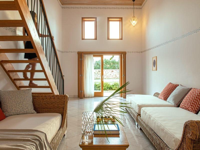 Soggiorno accogliente con divani in vimini, cuscini, scala in legno e grandi finestre con vista sul giardino.