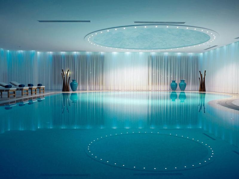 Piscina interna con illuminazione blu, sedie a sdraio, vasi e soffitto con lampada circolare.