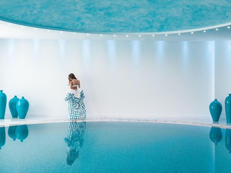 Donna in abito fluente accanto a vasi turchesi presso piscina interna con soffitto blu.