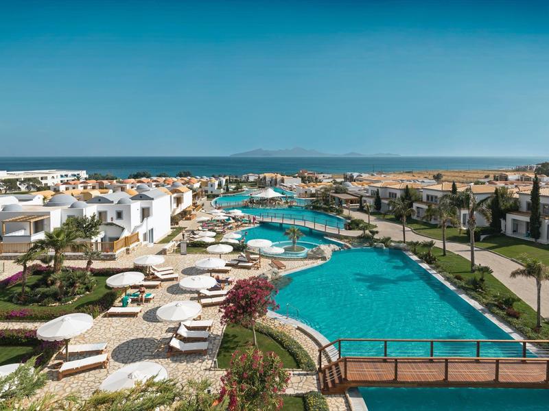 Area piscina di un hotel di lusso con edifici bianchi, ombrelloni e cielo azzurro.