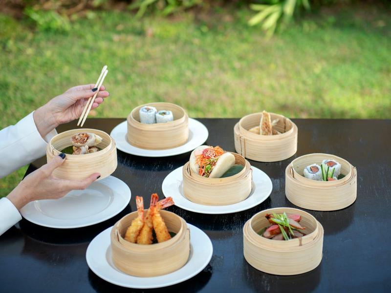 Mani che tengono un dumpling al vapore con bacchette accanto a vari piatti di dim sum in cestini di bambù.