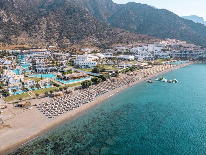 Resort sulla spiaggia con lettini e ombrelloni vicino al mare azzurro e montagne sullo sfondo.