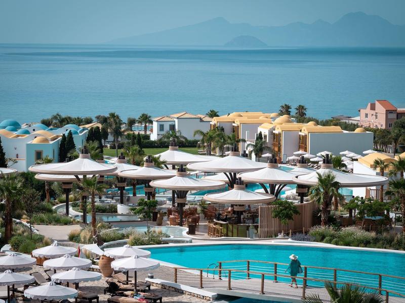 Area piscina del resort con ombrelloni e lettini con vista sul mare e montagne lontane.