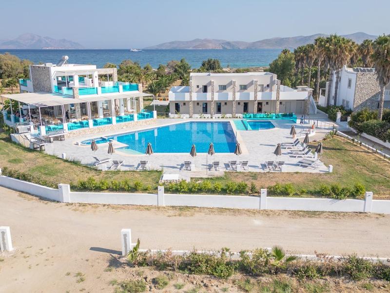 Widok na hotel z dużym odkrytym basenem, otoczony palmami i morzem w tle.