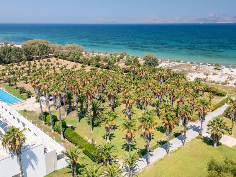 Widok na hotel z basenem, palmami i plażą przy morzu.