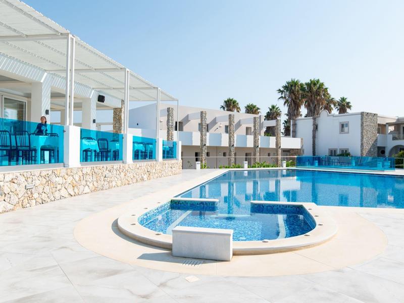 Nowoczesny basen hotelowy z niebieskimi leżakami i białymi budynkami w tle pod czystym niebem