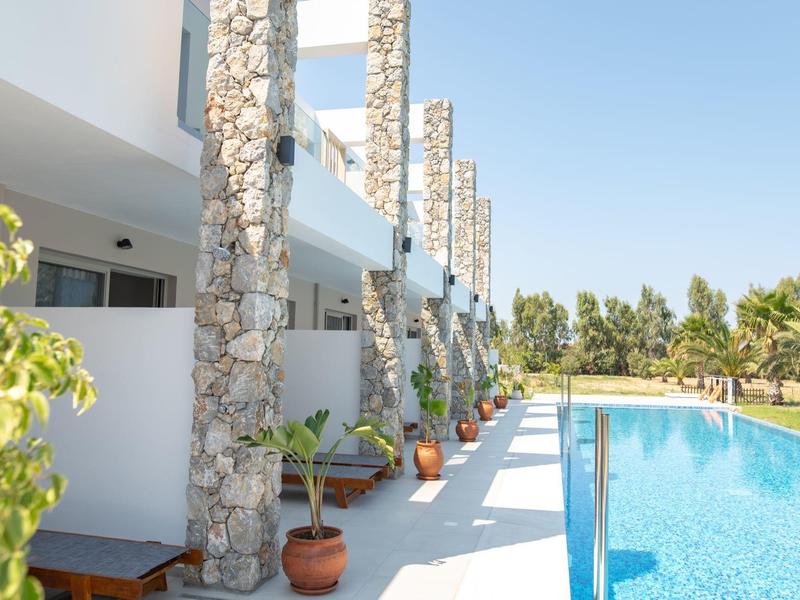 Zewnętrzny teren hotelu z basenem i kamiennymi filarami wzdłuż tarasu w słońcu.